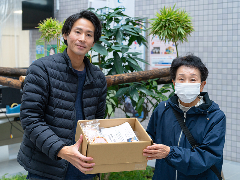 モカメルの中曽根雅也が、むつみ子ども食堂様にハニーワッフルが入った箱を寄付として手渡している様子