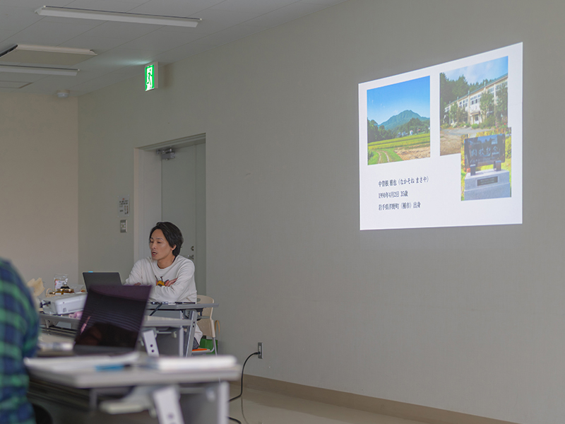 プロジェクターの前に座り、自身の経歴や活動について発表するモカメルの様子